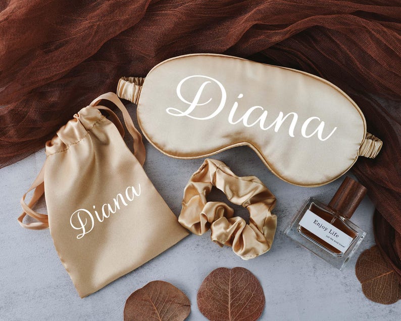 May include: A set of personalised gifts in a gold satin colour. The set includes a sleep mask, a small drawstring bag, a hair scrunchie, and a bottle of perfume. All items are personalised with the name "Diana".