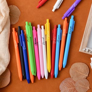 Puede incluir: Una colección de bolígrafos coloridos, cada uno con un nombre diferente impreso. Los bolígrafos están dispuestos sobre una superficie marrón, junto con un marco de madera y hojas decorativas. Los colores incluyen naranja, negro, azul, verde, rosa, blanco, amarillo y rojo.