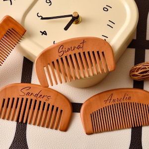 以下が含まれることがあります： 名前が刻まれた木製の櫛。「Simrat」、「Sanders」、そして「Aurelia」と書かれています。櫛は薄茶色で、細かい歯と湾曲したハンドルが付いています。長方形の櫛と時計も画像にあります。