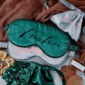 May include: Two satin sleep masks in green and grey with the names "Trisha" and "Sunny" written on them. The masks are on a brown and white fabric background.