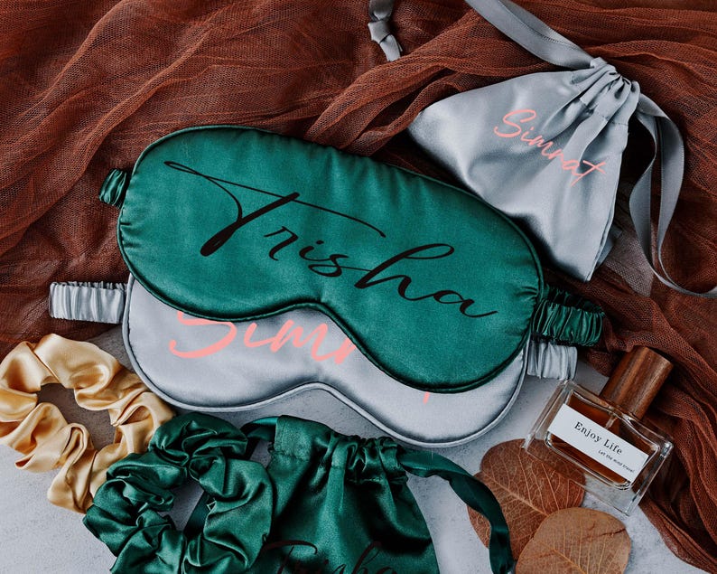 May include: Two satin sleep masks in green and grey with the names "Trisha" and "Sunny" written on them. The masks are on a brown and white fabric background.