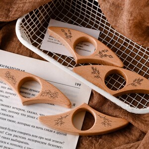 May include: Four wooden thumb page holders with a floral design are displayed. The holders are light brown and have a circular opening for the thumb. The holders are on a white wire basket and a book page.