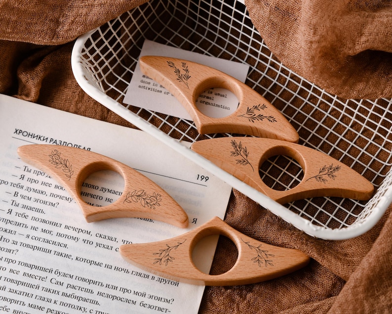 May include: Four wooden thumb page holders with a floral design are displayed. The holders are light brown and have a circular opening for the thumb. The holders are on a white wire basket and a book page.