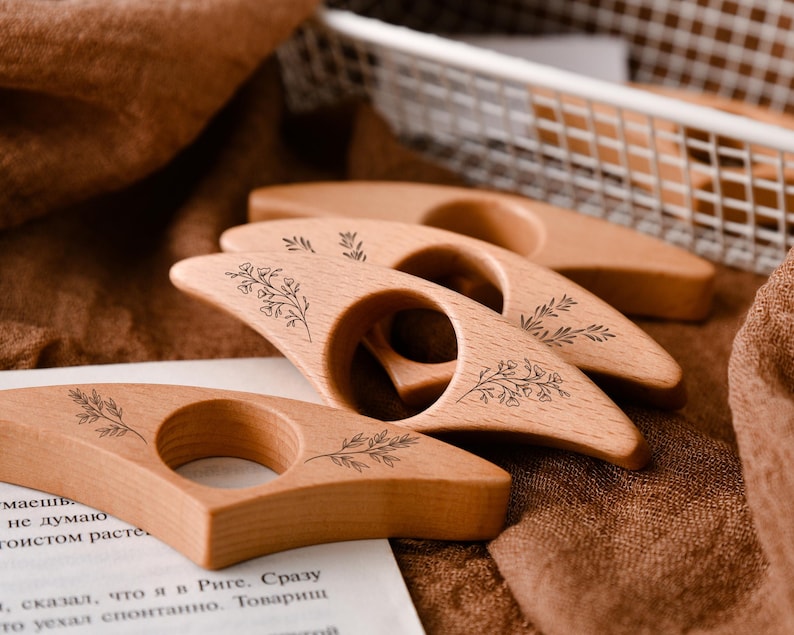 May include: Four wooden thumb page holders with floral designs are arranged on a brown surface. The light-colored wood has a smooth finish. A white basket and a piece of paper with text are in the background. The page holders are for books.