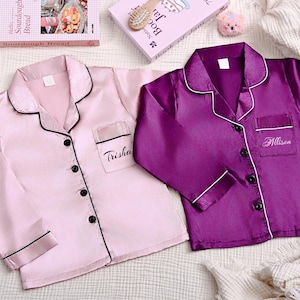 May include: Two silk pajama shirts, one light pink and one purple, are displayed. The pink shirt has the name "Trisha" embroidered on the pocket. The purple shirt has the name "Allison" embroidered on the pocket. Both shirts have black trim.