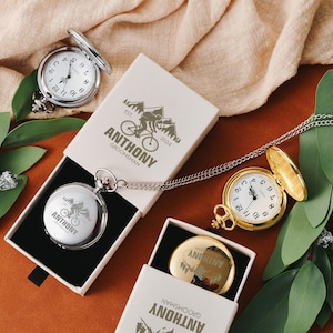 May include: Silver and gold pocket watches with white faces and black numerals, displayed open and closed. The watches are in white boxes with the name "ANTHONY GROOMSMAN" and a mountain bike graphic. The image is on a brown surface.