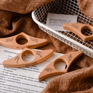 May include: Wooden thumb book page holders with floral designs and the word "Victoria" engraved on them. The book accessories are light brown and are resting on a book and a brown fabric.