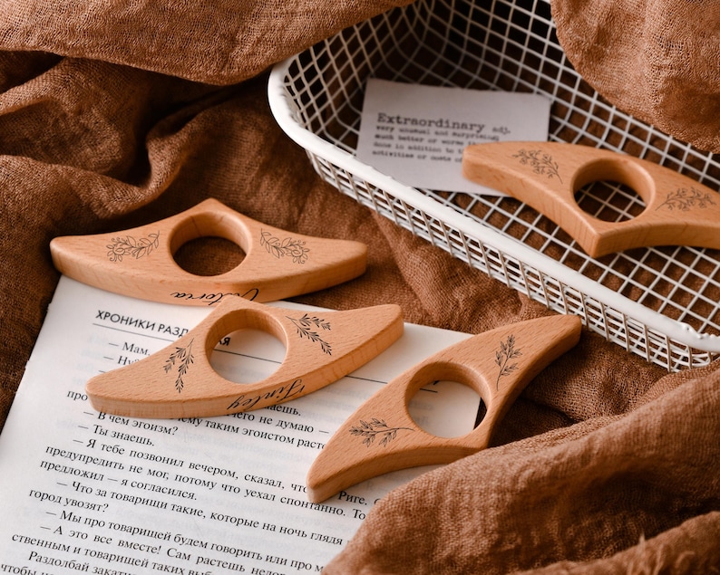 May include: Wooden thumb book page holders with floral designs and the word "Victoria" engraved on them. The book accessories are light brown and are resting on a book and a brown fabric.