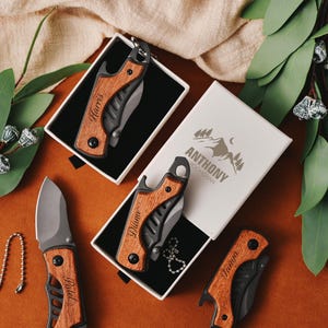 Puede incluir: Una colección de navajas de bolsillo personalizadas con mangos de madera y hojas negras. Algunas navajas están en cajas blancas, y una se muestra con una cadena. Las cajas tienen nombres grabados y una caja tiene el texto "ANTHONY SPORTSMAN".