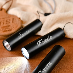 Può includere: Torce nere cilindriche con nomi incisi in corsivo. Le torce emettono una luce intensa. I nomi "Lauren", "Frieda" e "Sierra" sono visibili. Sullo sfondo, un'etichetta in pelle.