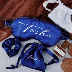 May include: A set of blue satin sleep accessories with the name "Trisha" embroidered on each piece. The set includes a sleep mask, a hair scrunchie, and a small pouch.