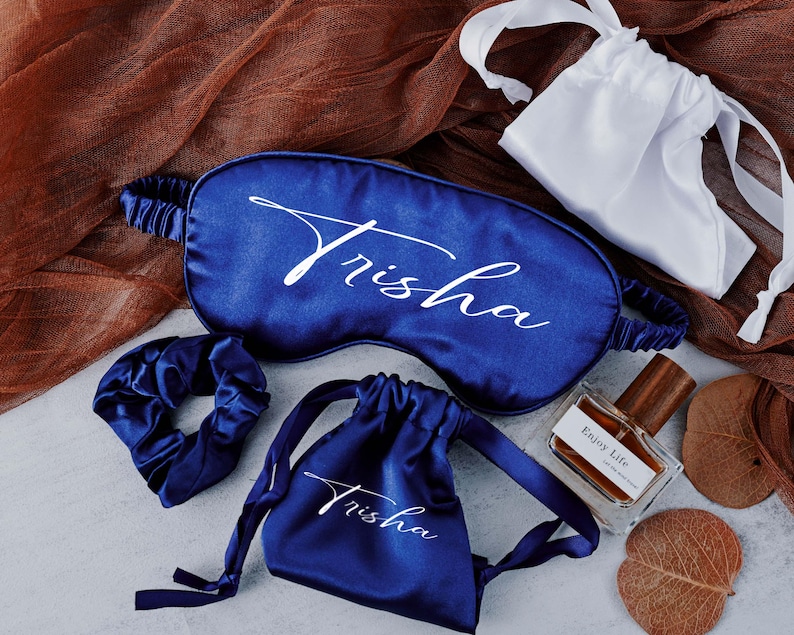 May include: A set of blue satin sleep accessories with the name "Trisha" embroidered on each piece. The set includes a sleep mask, a hair scrunchie, and a small pouch.