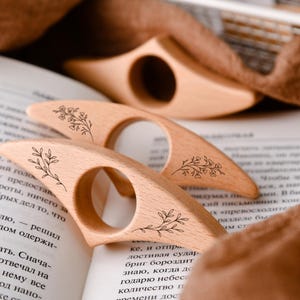 May include: Three wooden book page holders with floral designs are arranged on an open book. The light-colored wood has a smooth finish. The book page holders are designed to be held with a finger through the circular opening.