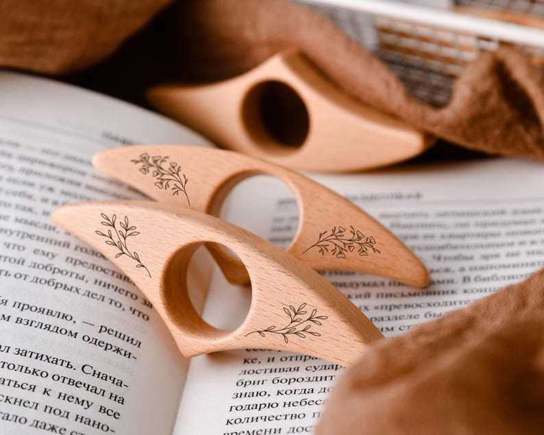 May include: Three wooden book page holders with floral designs are arranged on an open book. The light-colored wood has a smooth finish. The book page holders are designed to be held with a finger through the circular opening.