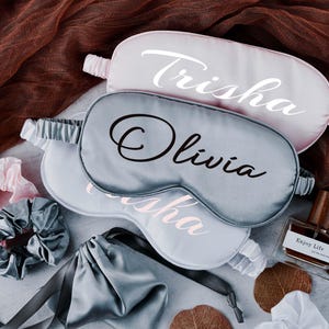 May include: Three satin sleep masks with personalised names: Trisha, Olivia, and Trisha. The masks are in shades of pink and grey.
