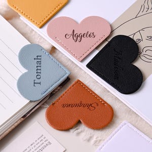 May include: A selection of heart-shaped leather bookmarks in colours such as yellow, pink, blue, black, and brown. Each bookmark has a name printed on it. The bookmarks are placed on open books and stationery.