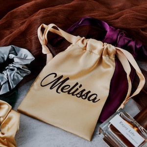 May include: A beige satin drawstring bag with the name "Melissa" printed in black. The bag is surrounded by two satin hair scrunchies, one in light blue and one in gold, and a purple satin ribbon.