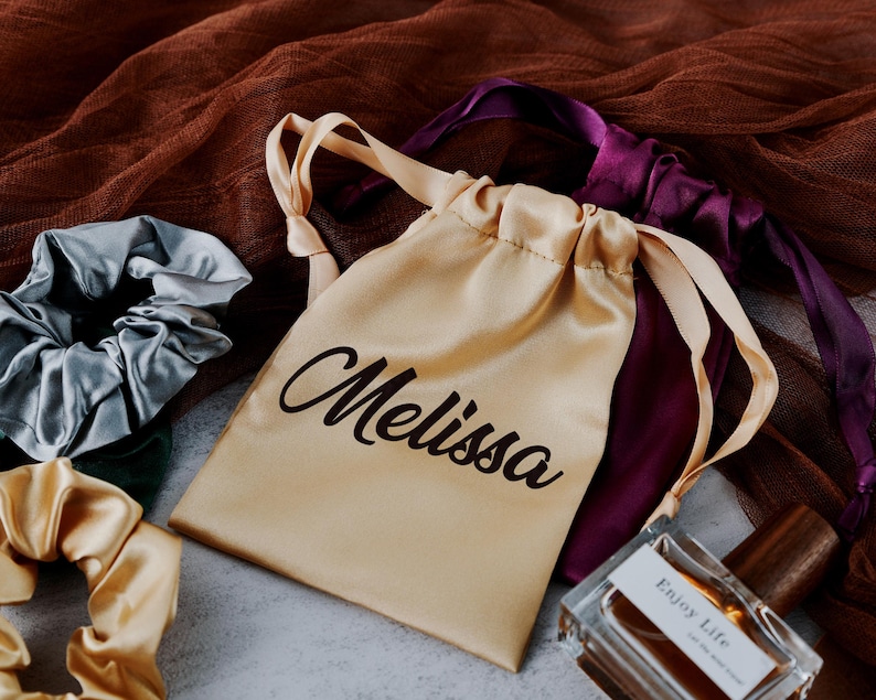 May include: A beige satin drawstring bag with the name "Melissa" printed in black. The bag is surrounded by two satin hair scrunchies, one in light blue and one in gold, and a purple satin ribbon.