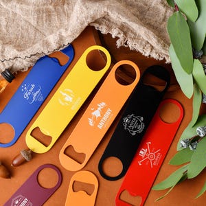 Può includere: Una collezione di apribottiglie colorati in blu, giallo, arancione, nero e rosso. Ogni apribottiglie ha un design e un testo diversi. Gli apribottiglie sono disposti su una superficie di legno con elementi decorativi.