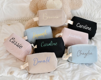 May include: A collection of personalized cosmetic bags in various colors, including pink, blue, black, and beige. Each bag features a name embroidered in a contrasting color, with names like "Cassie" and "Caroline". The bags have a textured appearance.