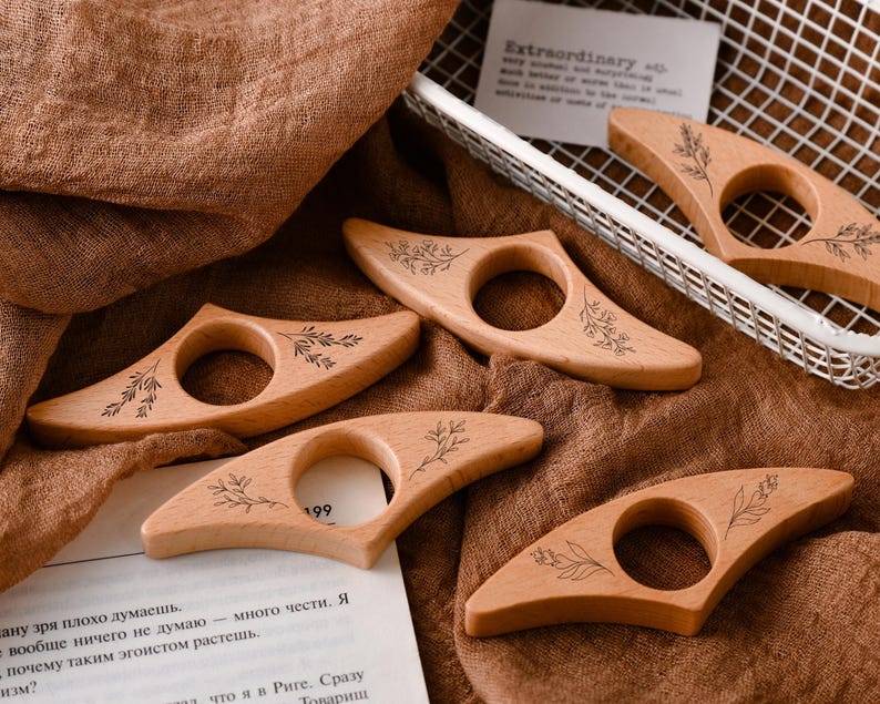 May include: Wooden thumb book holders with a floral design are displayed on a brown cloth. Each holder has a circular opening and a leaf pattern. The text "Extraordinary" is visible in the background.