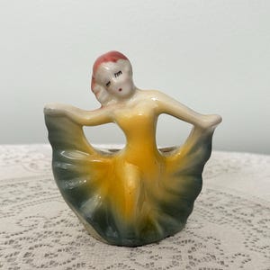May include: A vintage ceramic figurine of a dancer in a flowing dress. The figurine features a yellow bodice and a skirt that transitions from green to blue. The dancer has red hair and closed eyes, with arms outstretched.