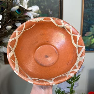 May include: A terracotta-colored bowl with a white geometric pattern around the rim. The bowl has a slightly textured surface and a small, circular indentation in the center. The bowl is likely handmade and has a rustic aesthetic.