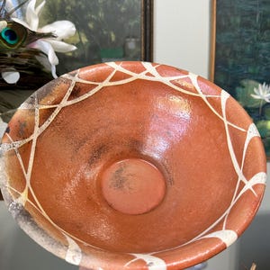 May include: A handmade ceramic bowl with a reddish-brown interior and a white geometric pattern around the rim. The bowl has a circular base and a wide opening, perfect for serving or display.