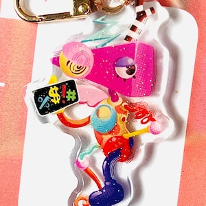 May include: A vibrant acrylic keyring with a gold-coloured clasp. The design showcases an abstract figure with pink, blue, yellow, and red elements. A small sign displays the symbols "%!$#".