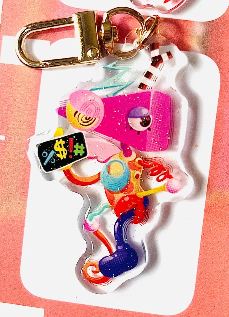 May include: A vibrant acrylic keyring with a gold-coloured clasp. The design showcases an abstract figure with pink, blue, yellow, and red elements. A small sign displays the symbols "%!$#".
