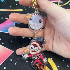 May include: A clear acrylic keyring with two cartoon characters. The top character is a winking, round creature with sharp teeth. The bottom character is a magician in a red coat and top hat. The keyring has a gold star and clasp.