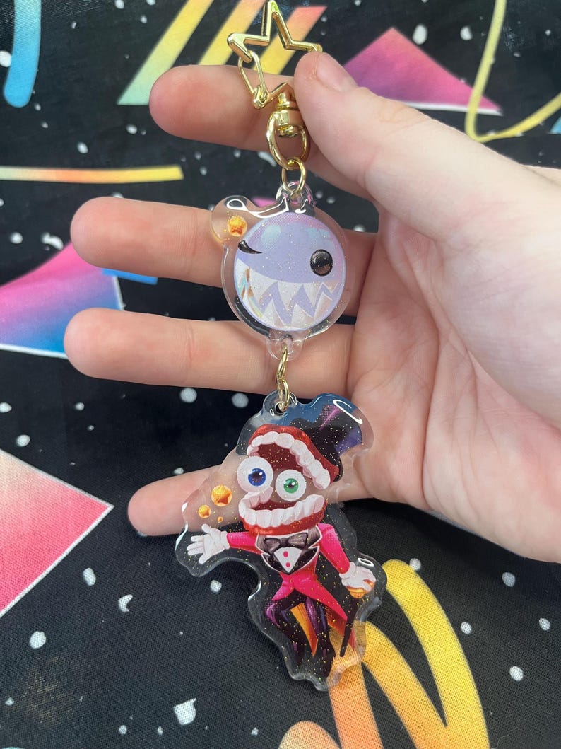 May include: A clear acrylic keyring with two cartoon characters. The top character is a winking, round creature with sharp teeth. The bottom character is a magician in a red coat and top hat. The keyring has a gold star and clasp.