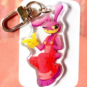 May include: A pink and red cartoon rabbit keyring with a gold-coloured clasp. The rabbit is wearing red dungarees and yellow gloves, winking and pointing. The keyring is on a white background with glitter accents.