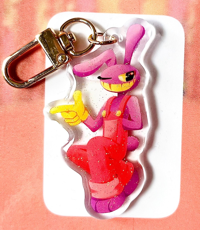 May include: A pink and red cartoon rabbit keyring with a gold-coloured clasp. The rabbit is wearing red dungarees and yellow gloves, winking and pointing. The keyring is on a white background with glitter accents.
