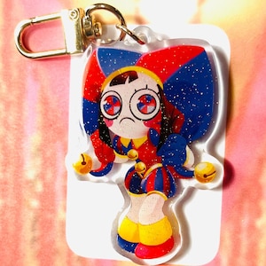 May include: A colourful acrylic keyring featuring a cartoon jester character with a sad expression. The jester has red, blue, and yellow clothing, with gold accents and a gold clasp. The keyring is approximately 7.6 cm in height.