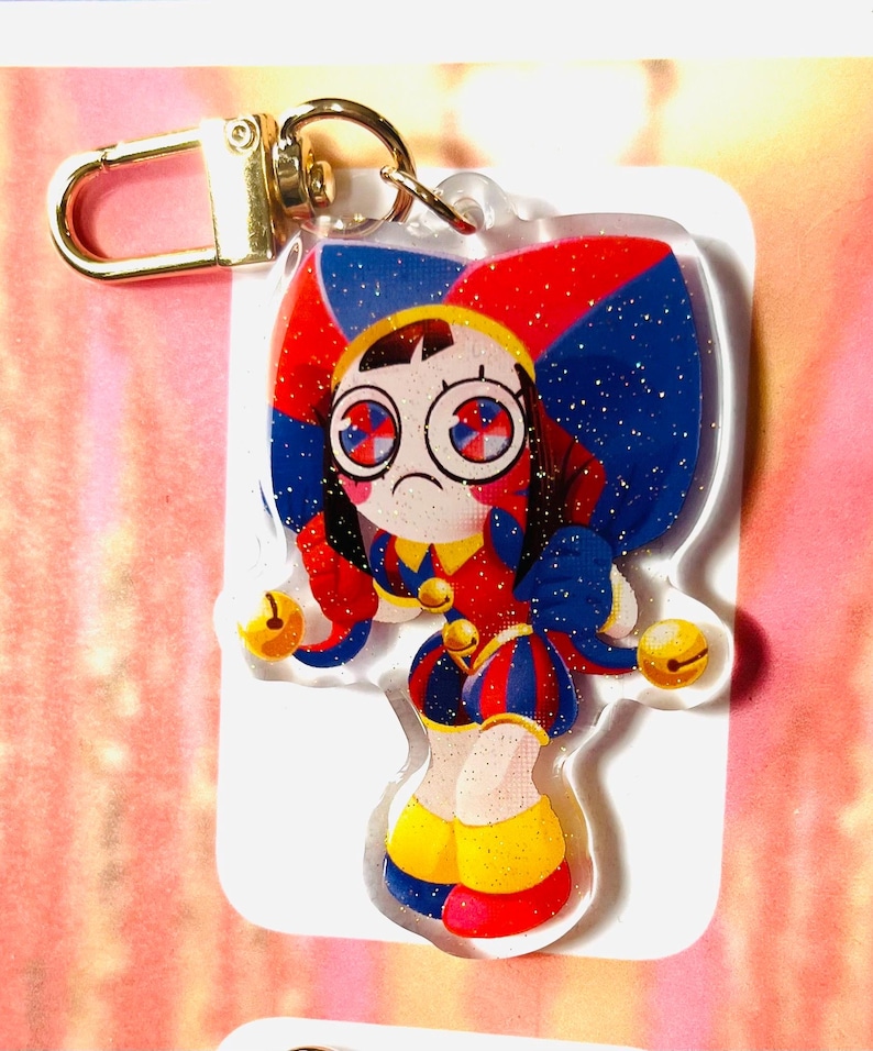 May include: A colourful acrylic keyring featuring a cartoon jester character with a sad expression. The jester has red, blue, and yellow clothing, with gold accents and a gold clasp. The keyring is approximately 7.6 cm in height.