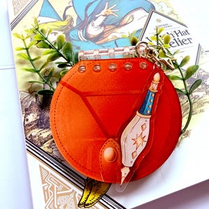 May include: A circular orange acrylic keychain featuring a stylized pen design with blue and gold accents. The keychain has a clear plastic spiral binding at the top and a small gold star-shaped charm attached to the metal ring, resting on a book.