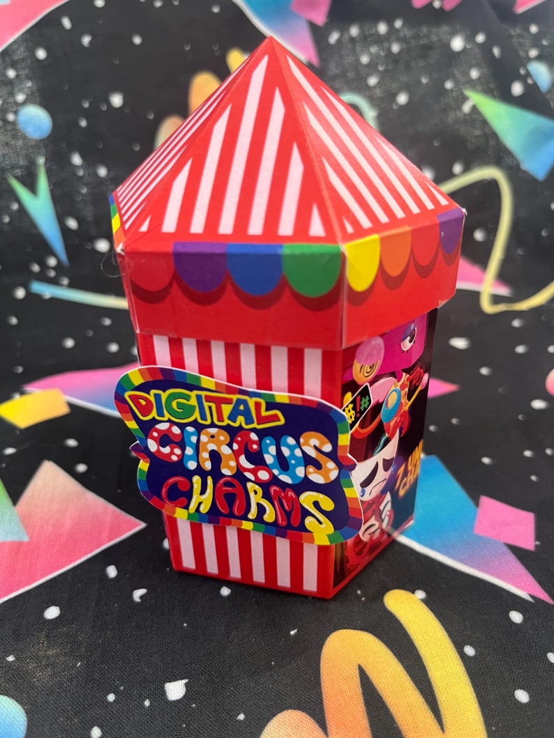 May include: A red and white striped circus-themed box with a rainbow-coloured trim. The box features the text "DIGITAL CIRCUS CHARMS" on a colourful sign. The background is a black fabric with colourful shapes.