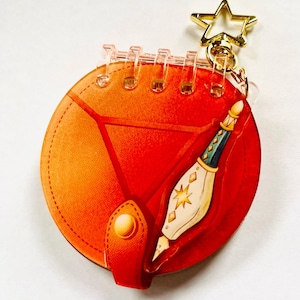 May include: A round, orange keychain resembling a notebook with a pen illustration. The keychain has a gold star-shaped clasp and clear plastic binding. The pen illustration is white, teal, and gold.