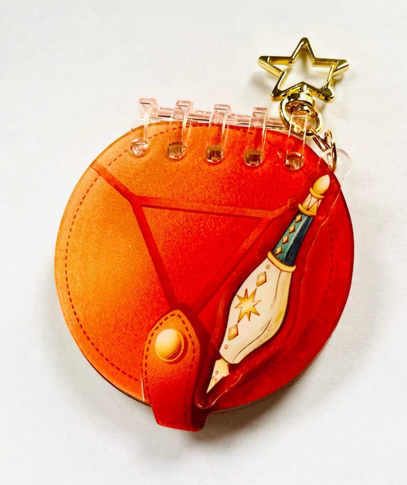 May include: A circular acrylic keychain shaped like a small notebook with a vibrant orange cover, a gold star-shaped clasp, and a printed illustration of a white and teal fountain pen with gold star accents on the front cover.