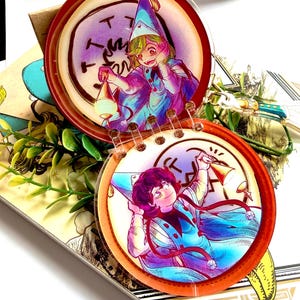 May include: A circular acrylic charm featuring two connected discs with illustrations of characters in pointed hats and capes. The top disc shows a character holding a lantern, while the bottom disc shows a character holding a bell, both with vibrant blue, purple, and orange color palettes.