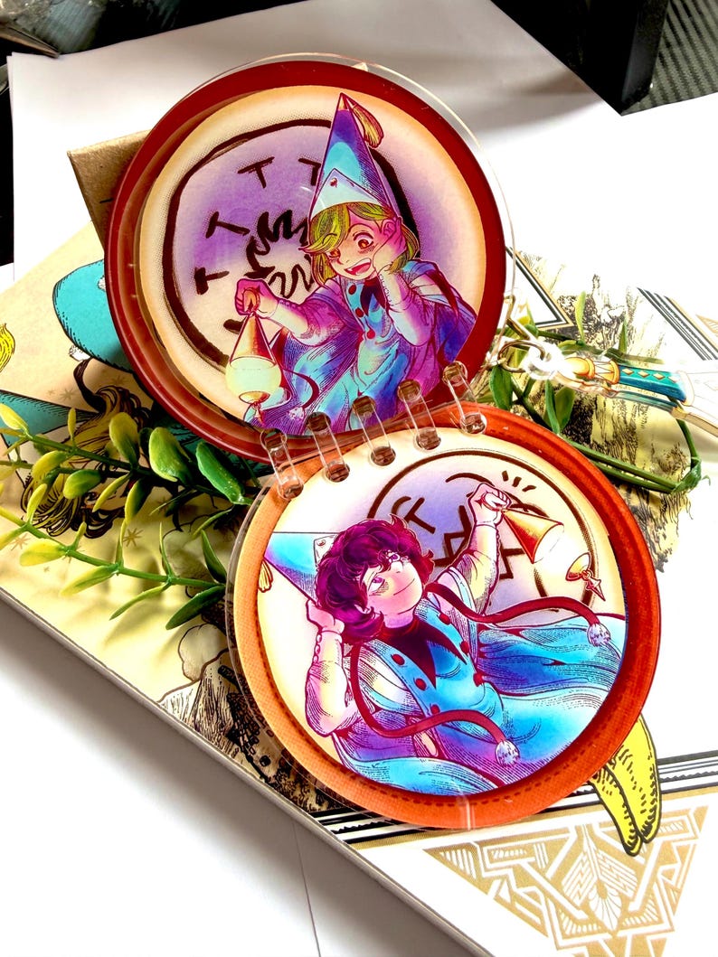 May include: A circular acrylic charm featuring two connected discs with illustrations of characters in pointed hats and capes. The top disc shows a character holding a lantern, while the bottom disc shows a character holding a bell, both with vibrant blue, purple, and orange color palettes.
