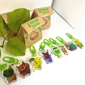 Könnte beinhalten: Eine Sammlung klarer Acryl-Schlüsselanhänger mit bunten Cartoon-Charakteren und grünen Verschlüssen. Die Schlüsselanhänger sind zusammen mit kleinen braunen Papiertüten mit der Aufschrift "KOROK SEEDS" angeordnet. Grüne Blätter sind ebenfalls vorhanden.