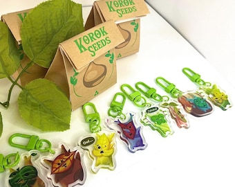 May include: A collection of clear acrylic keychains featuring colorful cartoon characters, with green clasps. The keychains are arranged with small brown paper bags labeled "KOROK SEEDS". Green leaves are placed near the bags.