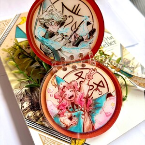 May include: A circular, hinged acrylic charm featuring two distinct illustrations of anime-style characters. The top disc shows a character with light hair and glasses, while the bottom disc depicts a character with pink hair making a peace sign.