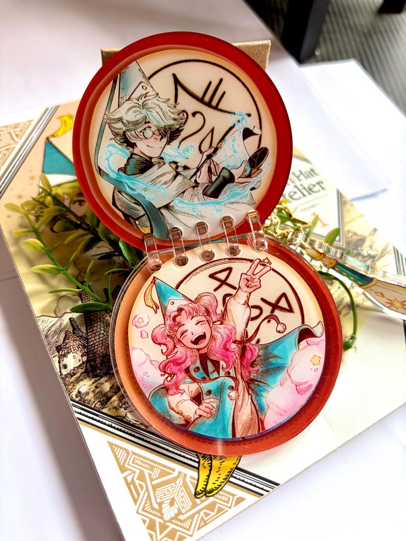 May include: A circular, hinged acrylic charm featuring two distinct illustrations of anime-style characters. The top disc shows a character with light hair and glasses, while the bottom disc depicts a character with pink hair making a peace sign.