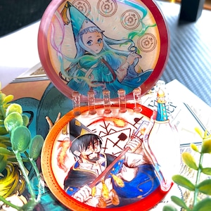 May include: A circular, hinged acrylic display piece featuring two distinct illustrations. The top disc shows a character in a blue pointed hat with magical symbols, while the bottom disc depicts a bearded character holding a staff with fire effects.