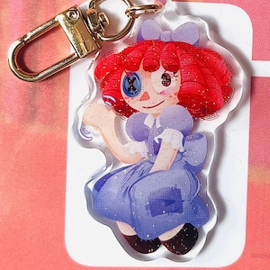 May include: A clear acrylic keyring featuring a cartoon doll with red hair, a blue dress, and a gold-coloured clasp. The doll has a button eye and a bow. The keyring is decorated with glitter.