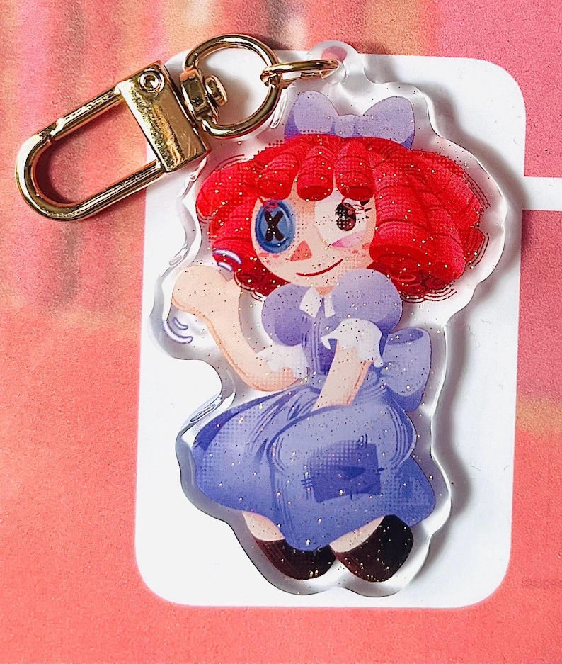 May include: A clear acrylic keyring featuring a cartoon doll with red hair, a blue dress, and a gold-coloured clasp. The doll has a button eye and a bow. The keyring is decorated with glitter.