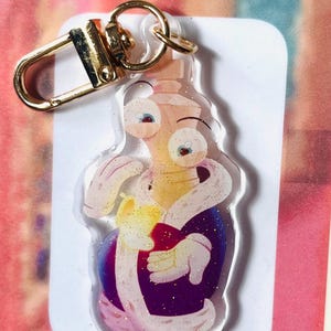 May include: A clear acrylic keyring featuring a cartoon character in a purple robe holding a red object. The keyring has a gold-coloured clasp and ring. The character has large eyes and is surrounded by glitter.